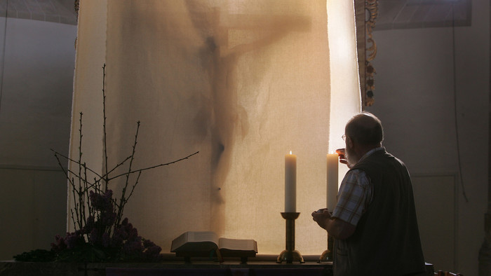 Mann in Weste zündet eine Kerze auf einem Altar an, im Hintergrund ist der Schatten von Jesus am Kreuz an der Wand.