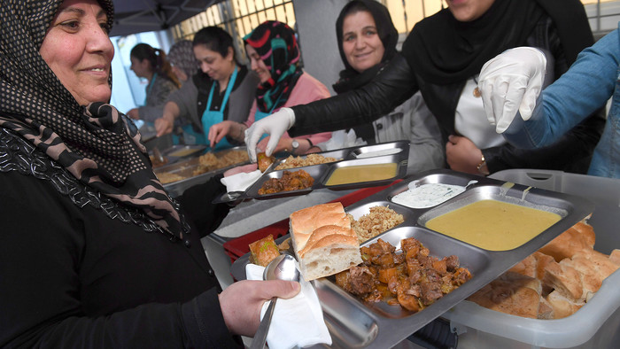 Frauen geben Essen aus, eine Frau im Vordergrund hält zwei Tablette mit mehreren Gerichten