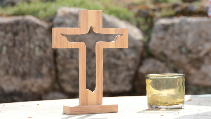 Kreuz aus Holf mit einer Aussparung, die Jesus am Kreuz erscheinen lässt. Daneben eine Kerze, beides draußen vor einer kleinen Steinmauer.
