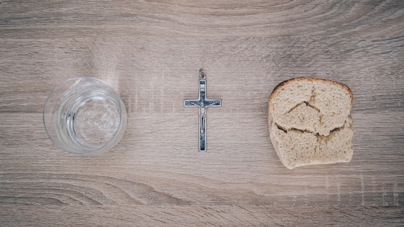 Auf einem Tisch stehen ein Glas Wasser, ein Kreuzanhänger mit Jesus und ein Stück Brot.