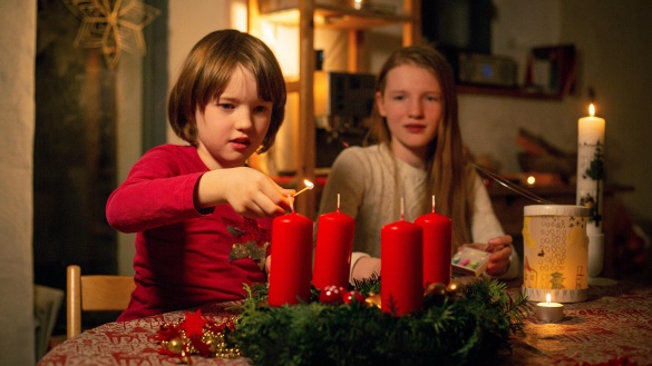 Mädchen und Junge mit grünem Adventskranz und roten, brennenden Kerzen.