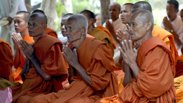 Ältere Mönche in orangenen Gewändern sitzen im Schneidersitz und haben ihre Hände vor ihrer Brust wie zum Gebet gefaltet