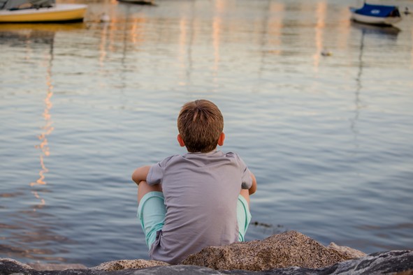 Ein Junge sitzt nachdenklich am Ufer und blickt aufs Wasser.