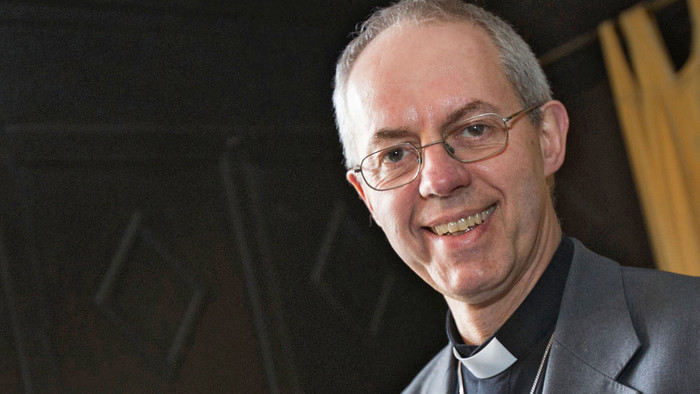 Bischof von Canterbury Justin Welby in grauem Jacket trägt ein großes silbernes Kreuz an einer Kette um den Hals. 