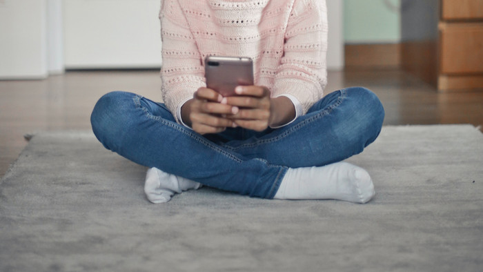 Kind in rosa Pullover, blauen Jeans, weißen Socken sitzt mit einem Handy in den Händen im Schneidersitz auf einem grauen Teppich. Der Kopf ist nicht auf dem Bildausschnitt
