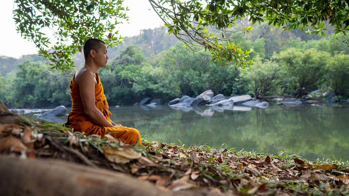 Ein junger Mann in orangenem Umhang meditiert im Schneidersitz an einem Fluss