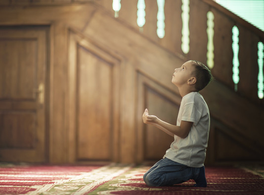 Ein muslimischer Junge betet auf Knien in einer Moschee mit offenen Händen und den Blick nach oben gerichtet.