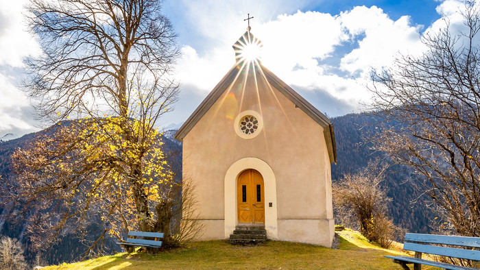 Außenansicht einer Kapelle im Grünen in den Bergen. Die Kapelle ist ein kleines helles Haus mit einer Holztür und einem spitzen Dach. Die Sonne strahlt im Hintergrund, über ihren Strahlen ist das Kreuz auf dem Dach der Kapelle