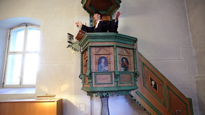 Ein Pfarrer mit ausgebreiteten Armen auf einer hohen Kanzel aus Holz und mit grünen Teilen und Bildern von Aposteln verziert
