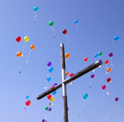 Holzkreuz vor blauem Himmel, um das bunte Luftballons fliegen
