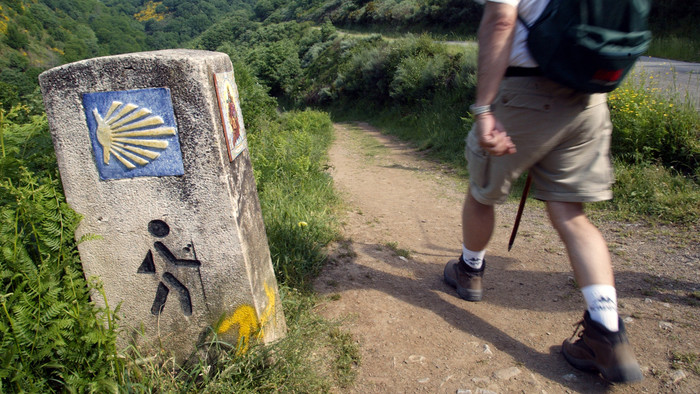 Hinweisschild Pilgerweg mit gelber Jakobsmuschel auf blauem Grund an einem Steinpfosten.