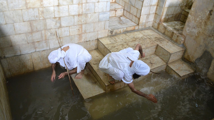 Zwei Männer in weißen Gewändern und Turbanen knieen auf hellen Steintreppen und beugen sich herunter zum Wasser