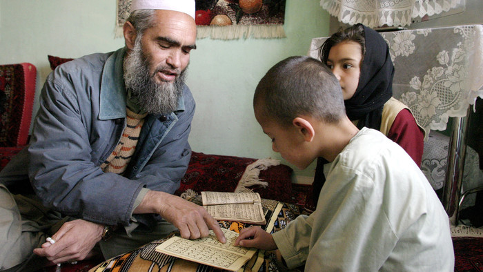 Ein älterer Mann zeigt auf einen Koranvers, ein Mädchen und ein Junge sitzen ihm gegenüber