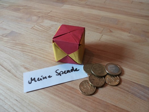Ein selbstgebasteltes Spendenkästchen aus rot-gelbem Papier, Kleingeld und ein Zettel mit der Aufschrift "Meine Spende".