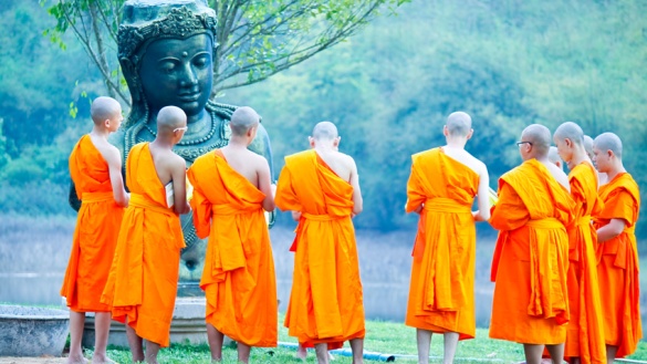 Buddhistische Mönche in orangefarbenen langen Wickelgewändern stehen auf einer Wiese, im Halbkreis um eine Buddhastatue