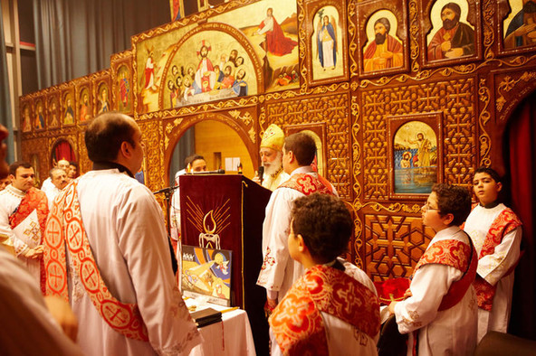 Golden verzierter Kirchenraum mit Bildern von Heiligen, im Vordergrund Männer und Jungs in weißen Gewändern mit roten Scherpen