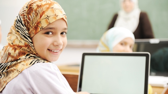 Ein muslimisches Mädchen im Klassenzimmer mit einem aufgeklappten Laptop vor sich.