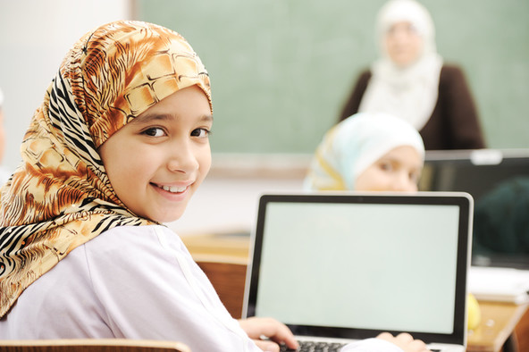 Ein Mädchen mit Kopftuch im Klassenzimmer mit einem aufgeklappten Laptop vor sich.