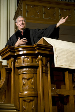 Ein evangelischer Pfarrer predigt in einer Kirche von einer Kanzel aus dunklem Holz zur Gemeinde.