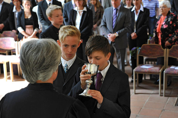 Zwei Jungen stehen bei einem Konfirmationsgottesdienst vor einem Pfarrer und einer der Jungen trinkt aus einem goldenen Kelch.