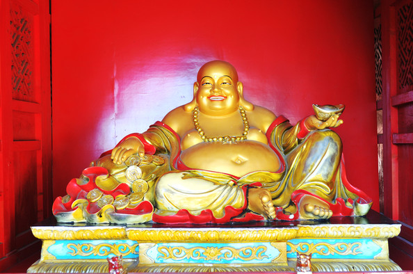 Lachender goldener Maitreya Buddha vor rotem Hintergrund mit einem Wassergefäß in seiner linken Hand.