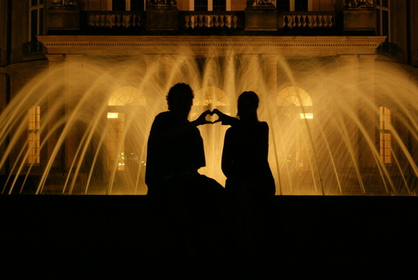 Silhouette eines Paares vor einem beleuchteten Brunnen bei Nacht, die mit den Händen ein Herz formen.