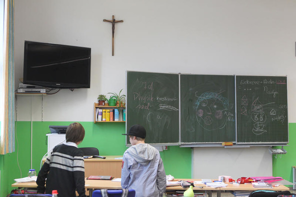 Zwei Jungen stehen in einem Klassenraum. Über der Tafel hängt ein Kreuz an der Wand.