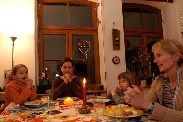 Zwei Mädchen sitzen mit gefalteten Händen an einem gedeckten Esstisch, auf dem eine Kerze brennt.