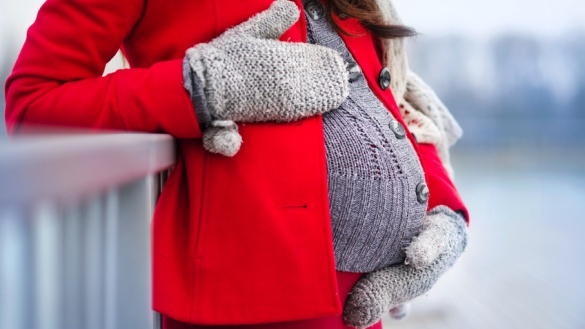 Eine schwangere Frau mit einer roten Jacke umfasst ihren Bauch mit beiden Händen. 