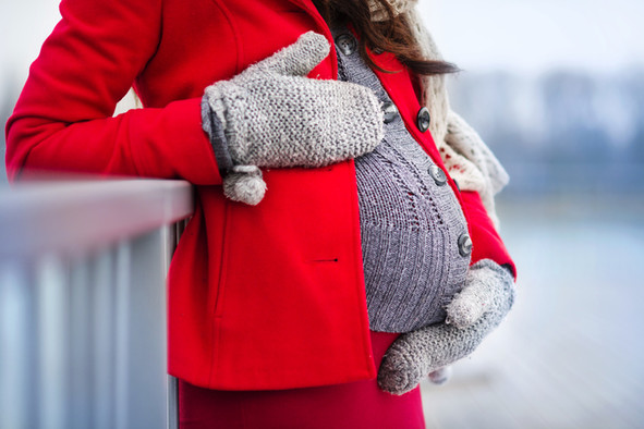 Eine schwangere Frau mit einer roten Jacke umfasst ihren Bauch mit beiden Händen. 