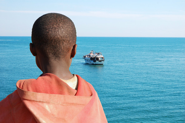 Ein Junge ist von hinten zu sehen und blickt auf ein Boot draußen auf dem Meer.