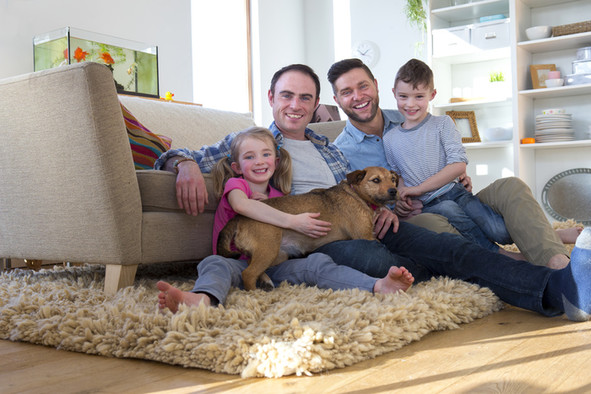 Ein homosexuelles männliches Paar sitzt mit Tochter, Sohn und Hund auf einem Teppich.