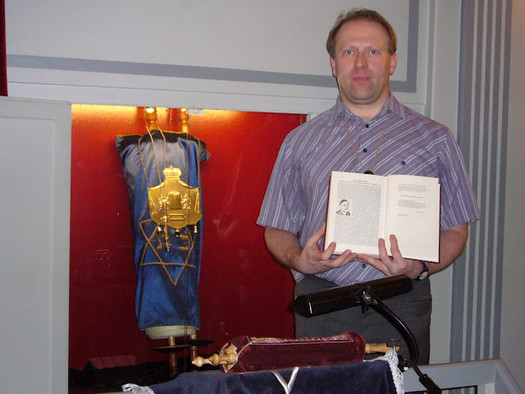 Eine Torarolle in einem Toraschrein, der mit rotem Samt ausgkleidet ist. Daneben steht ein Mann und hält ein aufgeschlagenes Buch vor sich.