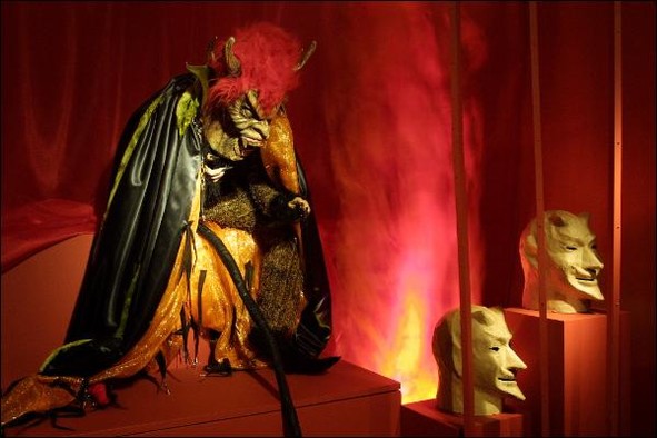 Eine Teufelsgestalt mit schwarzem Umhang, langem Schwanz, Hörnern und roten Haaren in einer Ausstellung.