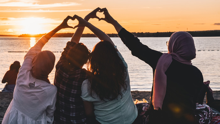 Vier junge Frauen sitzen an einem Strand mit Sonnenuntergang und halten jeweil eine Hand zur Hand einer anderen so, dass Herzen gebildet werden.