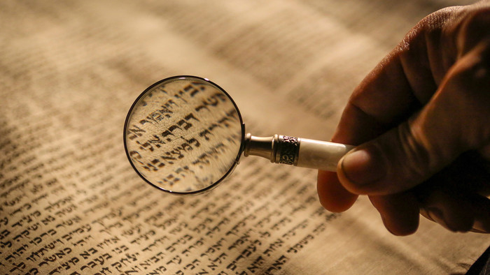 Hand hält kleine Lupe über einen alt erscheinenden Text, durch die Lupe sind hebräische Schriftzeichen erkennbar
