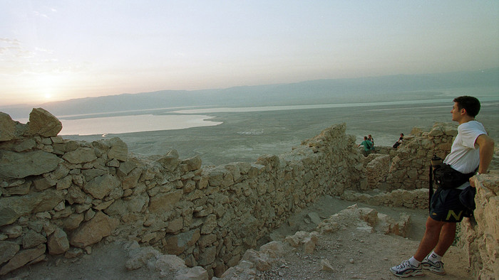 Ein Mann in weißem T-Shirt und kurzer Shorts steht zwischen alten, hellen Mauern, die zum Teil beschädigt sind und blickt Richtung Sonnenaufgang über das tote Meer.