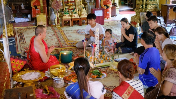 Ein Mönch und weitere Menschen sitzen auf dem Boden eines Tempels mit betenden Händen.