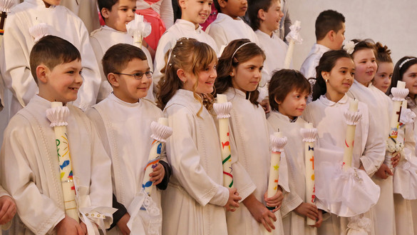 Kinder in weißen Gewändern halten ihre Taufkerzen und lächeln.