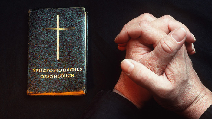 Links ein schwarzes Neuapostolisches Gesangbuch mit Schrift und Kreuz in gold, rechts zwei Hände zum Gebet gefaltet