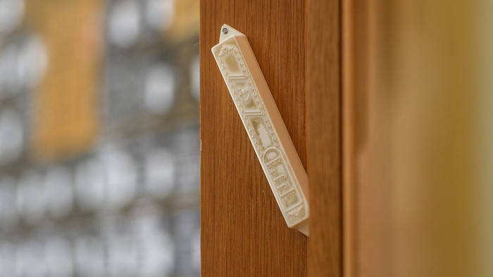 Eine Kapsel, in der der eine Pergamentrolle steckt, hängt in einem Türrahmen.