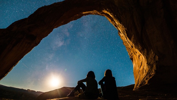zwei Menschen sitzen in einer felsigen Landschaft bei Nacht und schauen in den Sternenhimmel