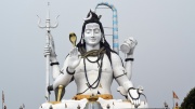 Große weiße Statue von Shiva vor grauem Himmel. In einer Hand hält er einen Dreizack, in einer eine Trommel und mit zwei Händen zeigt er Gesten. Eine Schlange ist um seinen Hals gewunden