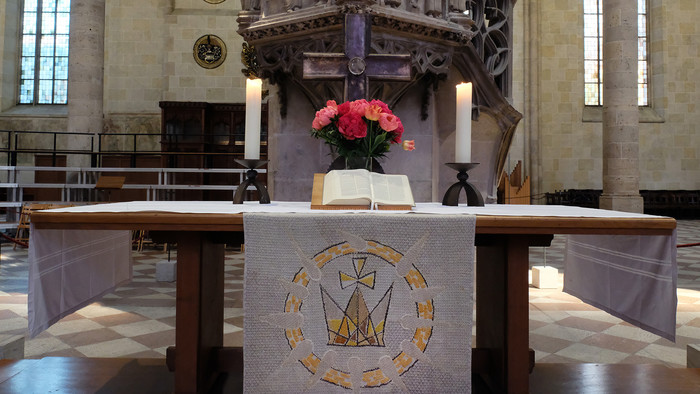 Altartisch aus Holz, bedeckt mit weißen Tüchern, die wiederum mit einer goldenen Krone verziert ist. Auf dem Tisch stehen zwei brennende Kerzen, dazwischen ein Kreuz, eine Vase mit roten und rosa Blumen und davor eine aufgeschlagene Bibel.