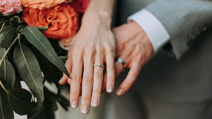 Die Hand einer Braut und eines Bräutigams mit Eheringen liegen aufeinander. Darunter ist ein Brautstrauß zu sehen.