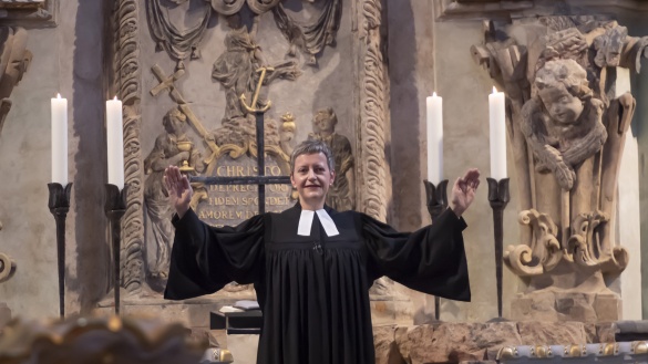 Eine Pfarrerin hält bei einem Gottesdienst vor einem Altar die segnenden Hände ausgebreitet nach oben.