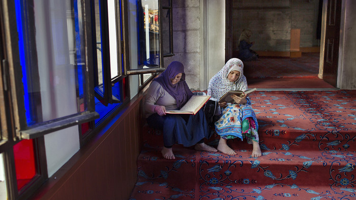 Zwei Frauen sitzen auf den bunten Stufen in einer Moschee und lesen jeweils im Koran. Im Hintergrund betet eine Frau im Moscheeraum.