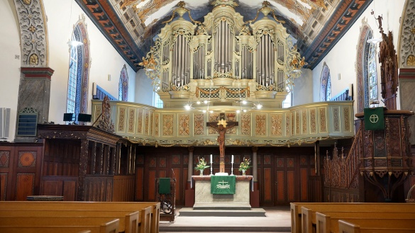 Ein großer verzierter Kirchen-Innenraum mit Altar, Kreuz und heller Orgel.
