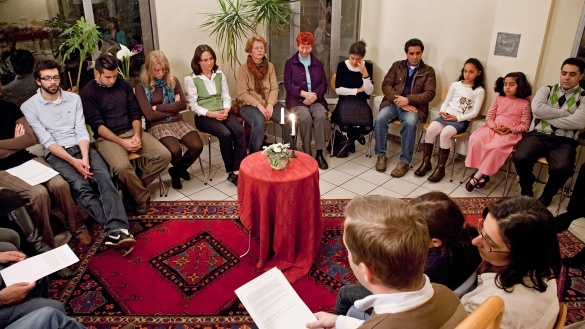 Menschen verschiedenen Alters sitzen auf Stühlen in einem Kreis um einen kleinen Tisch herum. Auf dem Tisch liegt eine rote Tischdecke und zwei Kerzen brennen. Manche Personen halten einen Zettel in der Hand.