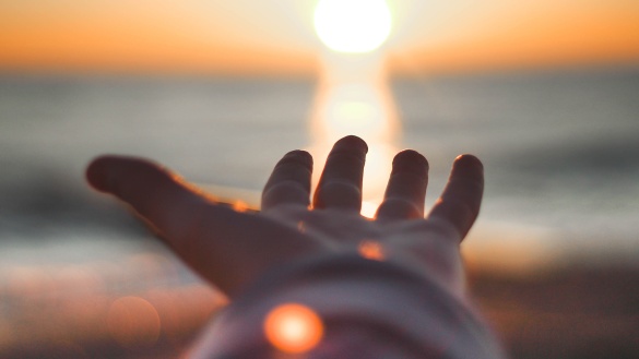 Eine Person am Meer hält die Hände hoch zur untergehenden Sonne.